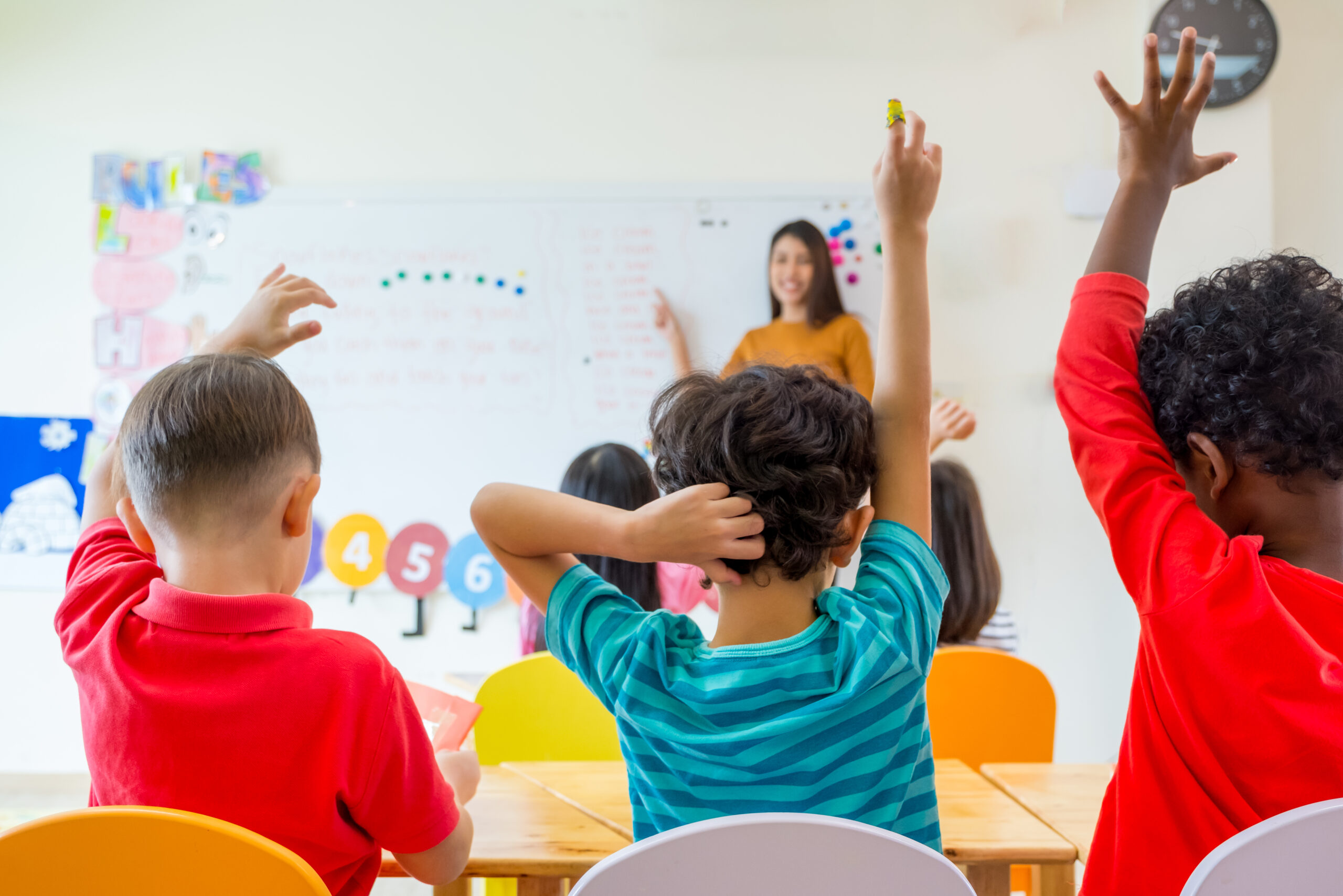 A teacher stands before a classroom of children raising their hands.